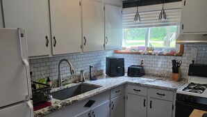 Una cocina con armarios blancos, un refrigerador, un lavaplatos, una estufa y una ventana con vistas a la vegetación.