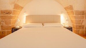 Un llit amb capçalera blanca i dues taules de nit amb làmpades, davant d'una paret de pedra amb sostre apuntat.