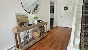A wooden console table with decorative items, a mirror, and a painting in a hallway with wooden flooring and a staircase.