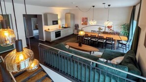 Een moderne woonkamer met een groene bank, een houten salontafel en hangende hanglampen. In de achtergrond is een keuken met witte kasten en een eethoek met een houten tafel en stoelen zichtbaar.