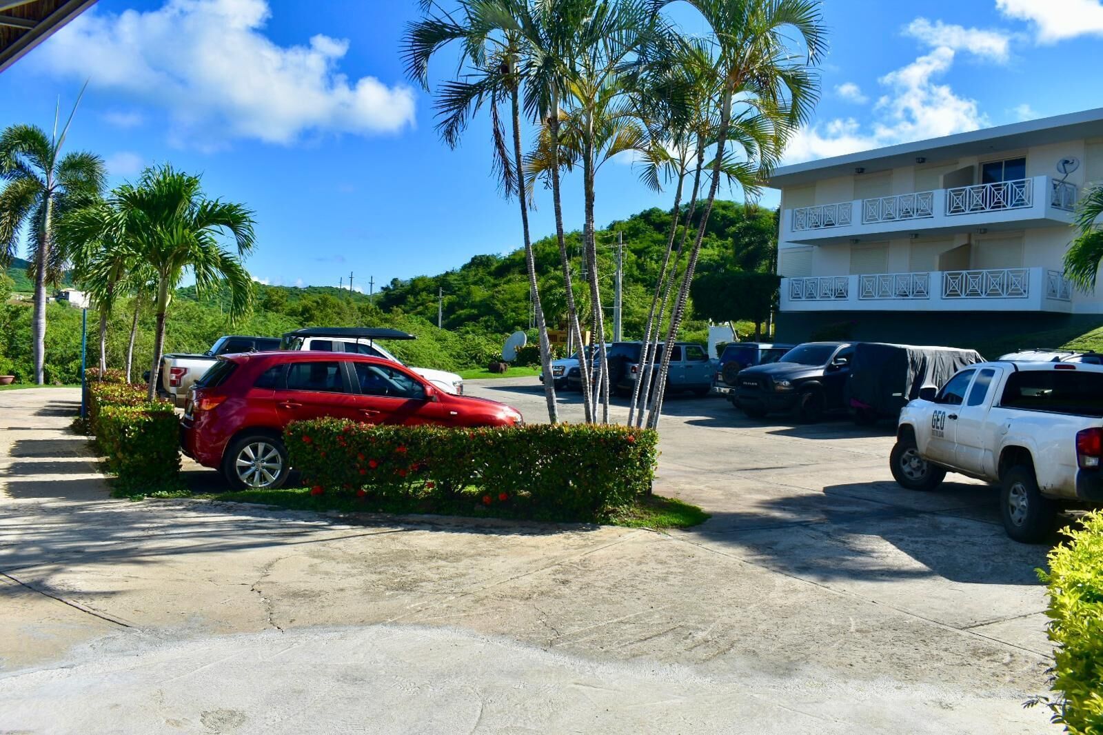 Un estacionamiento con varios coches, incluyendo un SUV rojo y una camioneta blanca, rodeado de palmeras y un edificio con balcones.