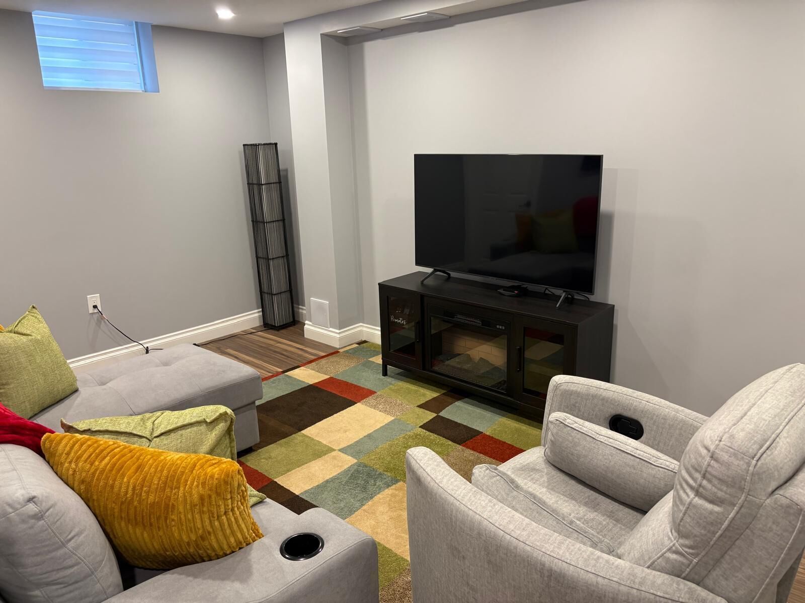 Una sala de estar con un televisor de pantalla plana sobre un soporte negro, sofás grises, un tapete colorido y una estantería alta de color negro.