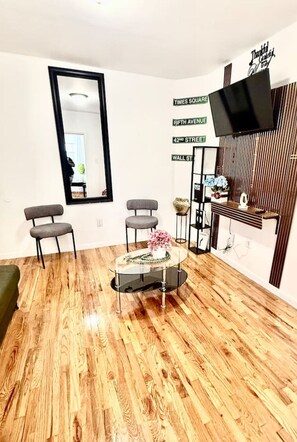 Ein Zimmer mit Holzboden, zwei graue Stühle, einem runden Glastisch mit einer Vase, einem großen Spiegel und einem an der Wand befestigten Fernseher.