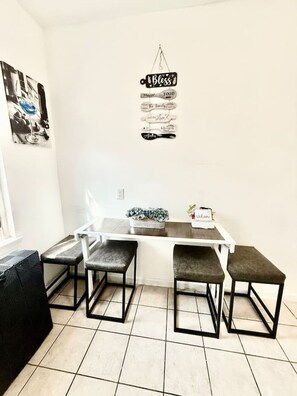 A small dining area with a white table and four grey stools. Above the table, there is a sign with the word "Bless" and other text.