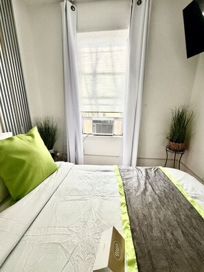 Ein Schlafzimmer mit Bett, Fenster mit Vorhängen, einer Zimmerpflanze und einem an der Wand befestigten Fernseher.
