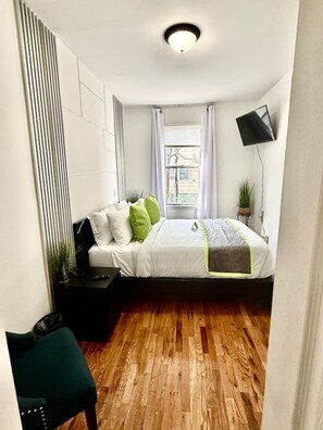 A modern bedroom with a bed, a bedside table, a chair, a potted plant, and a window with curtains.