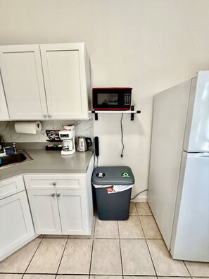 Eine kleine Küche mit weißen Schränken, einer Mikrowelle, einer Kaffeemaschine, einem Mülleimer und einem Kühlschrank.