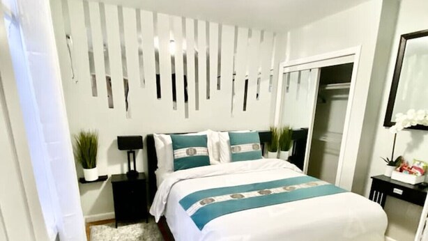 A bedroom with a bed, bedside tables, a nightstand, a mirror, a door, and a wall-mounted light fixture.
