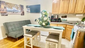 A compact kitchen with wooden cabinets, a dining table with two stools, and a grey sofa.