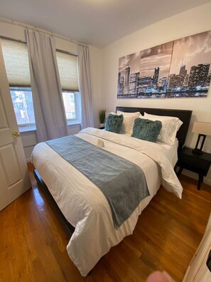 A bedroom with a bed, a nightstand, a window with curtains, and a cityscape painting on the wall.