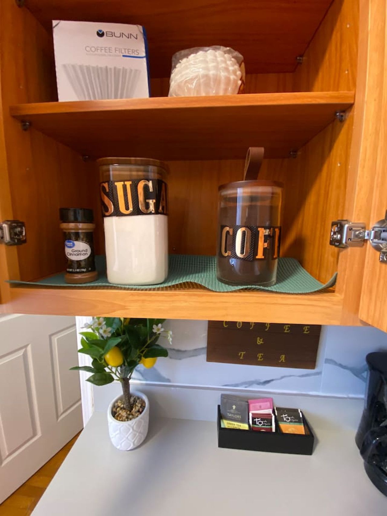A kitchen shelf with a box of Bunn coffee filters, a jar labeled 'SUGAR', a jar labeled 'COFFEE', and a small plant. Below, there's a coffee maker and a tray with tea bags.
