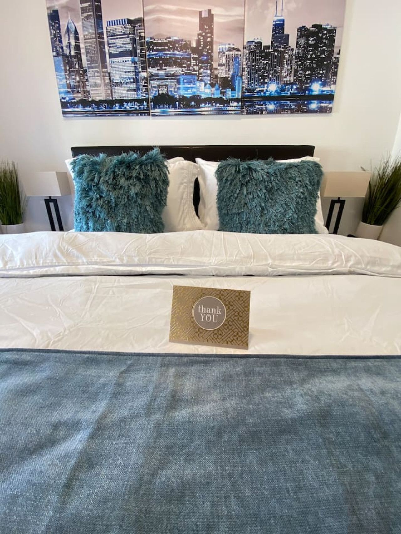 A neatly made bed with fluffy pillows, a thank you card, and a cityscape painting above.