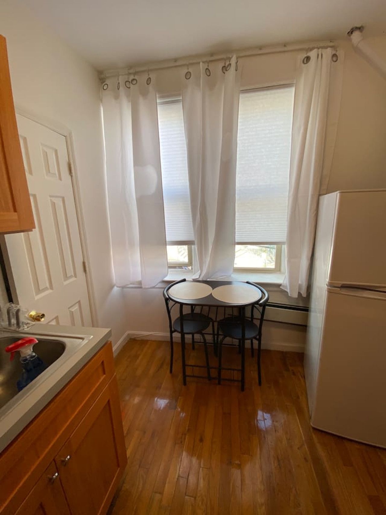 A small kitchen with a refrigerator, a sink, and a window with curtains. There is a small table with two chairs.