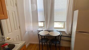 A small kitchen with a refrigerator, a sink, and a window with curtains. There is a small table with two chairs.