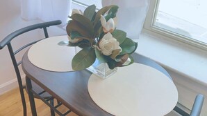 A round dining table with two chairs, a vase with flowers, and a window with curtains. - Comfy Getaway Private Apt Studio (West New York)