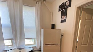 A compact kitchen with a refrigerator, a dining table with chairs, and a window with curtains. - Comfy Getaway Private Apt Studio (West New York)