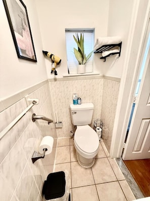 A small bathroom with a toilet, a towel rack, a window with a plant, and a framed picture on the wall.