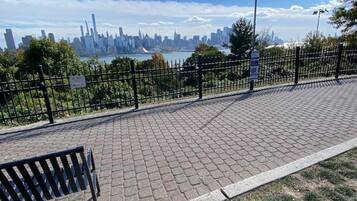 A paved walkway with a bench, a metal fence, and a city skyline in the background.