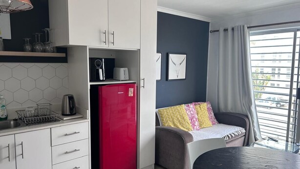 A compact kitchenette with a red refrigerator, a coffee maker, and a kettle. There is a dining area with a table and a sofa with patterned cushions. - Modern Sea Side Studio, a Surfer, Beach Goer or Cape Town Explorer's Paradise (Cape Town)