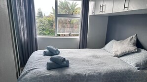 A neatly made bed with a patterned bedspread, a pillow, and rolled towels on a wooden floor. - Modern Sea Side Studio, a Surfer, Beach Goer or Cape Town Explorer's Paradise (Cape Town)