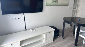 A room with a flat-screen TV mounted on the wall, a white bench, a round table, and a framed picture of an elephant. - Modern Sea Side Studio, a Surfer, Beach Goer or Cape Town Explorer's Paradise (Cape Town)