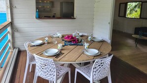 A round wooden table set for four with white wicker chairs, a small window above, and a view of the pool area. - KAY BLUE Creole house in the heart of nature, 2 steps from the beaches, magnificent (Les Trois-Îlets)