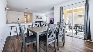 A dining area with a wooden table and chairs, a kitchen in the background, and a sliding glass door leading to an outdoor patio. - New Build Close to River & Golf! (Fort Mohave)