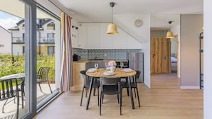 A modern kitchen with a dining area, featuring a wooden table set for four, a refrigerator, and a view of the outdoors. - Apartamenty Sun & Snow Bałtycka Oaza (Wladyslawowo)