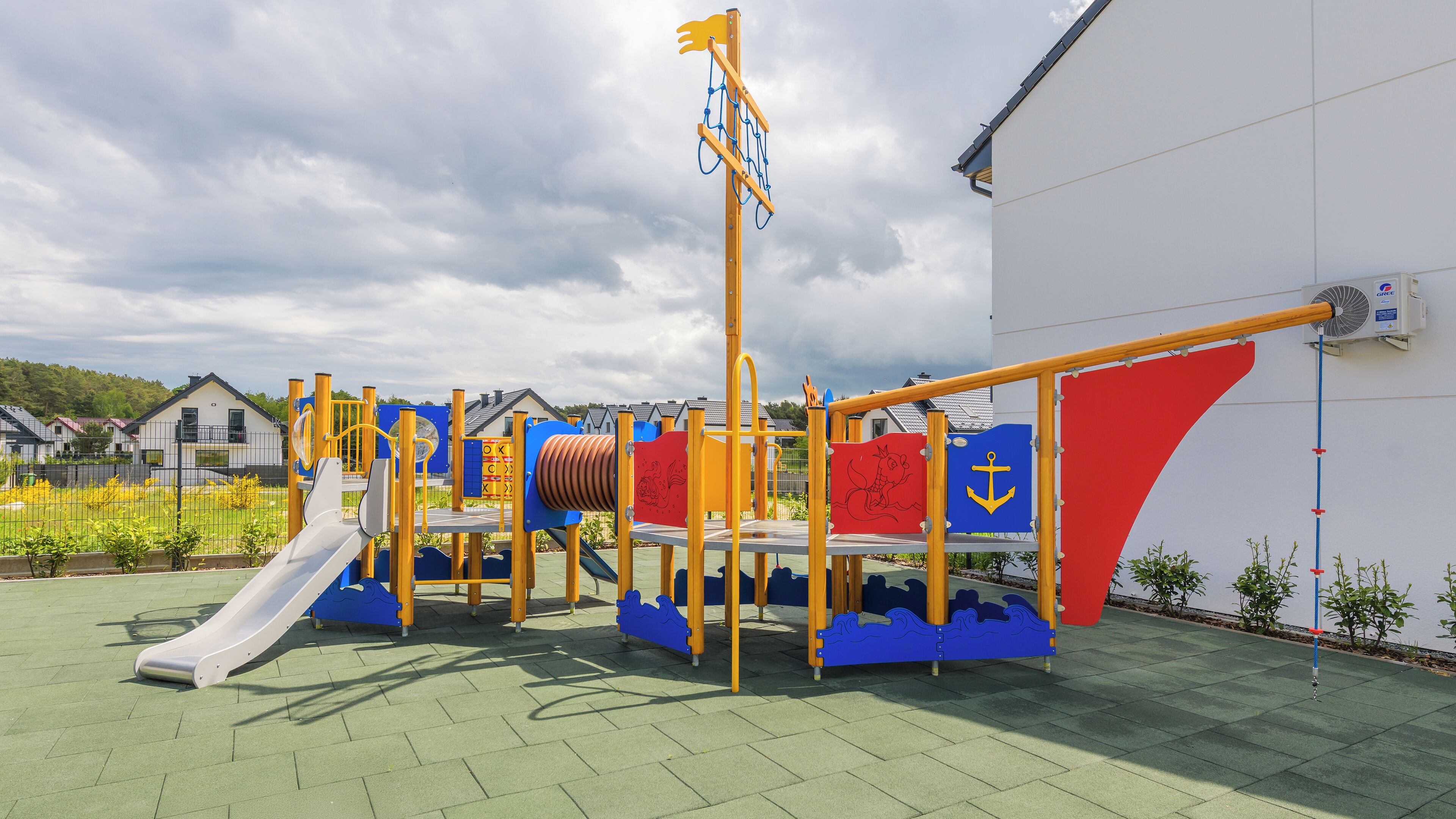 A playground with various play structures, including a slide and climbing elements, set against a backdrop of residential houses and a white building.