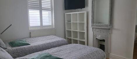 A bedroom with two beds, a white radiator, a TV mounted on the wall, and a fireplace.
