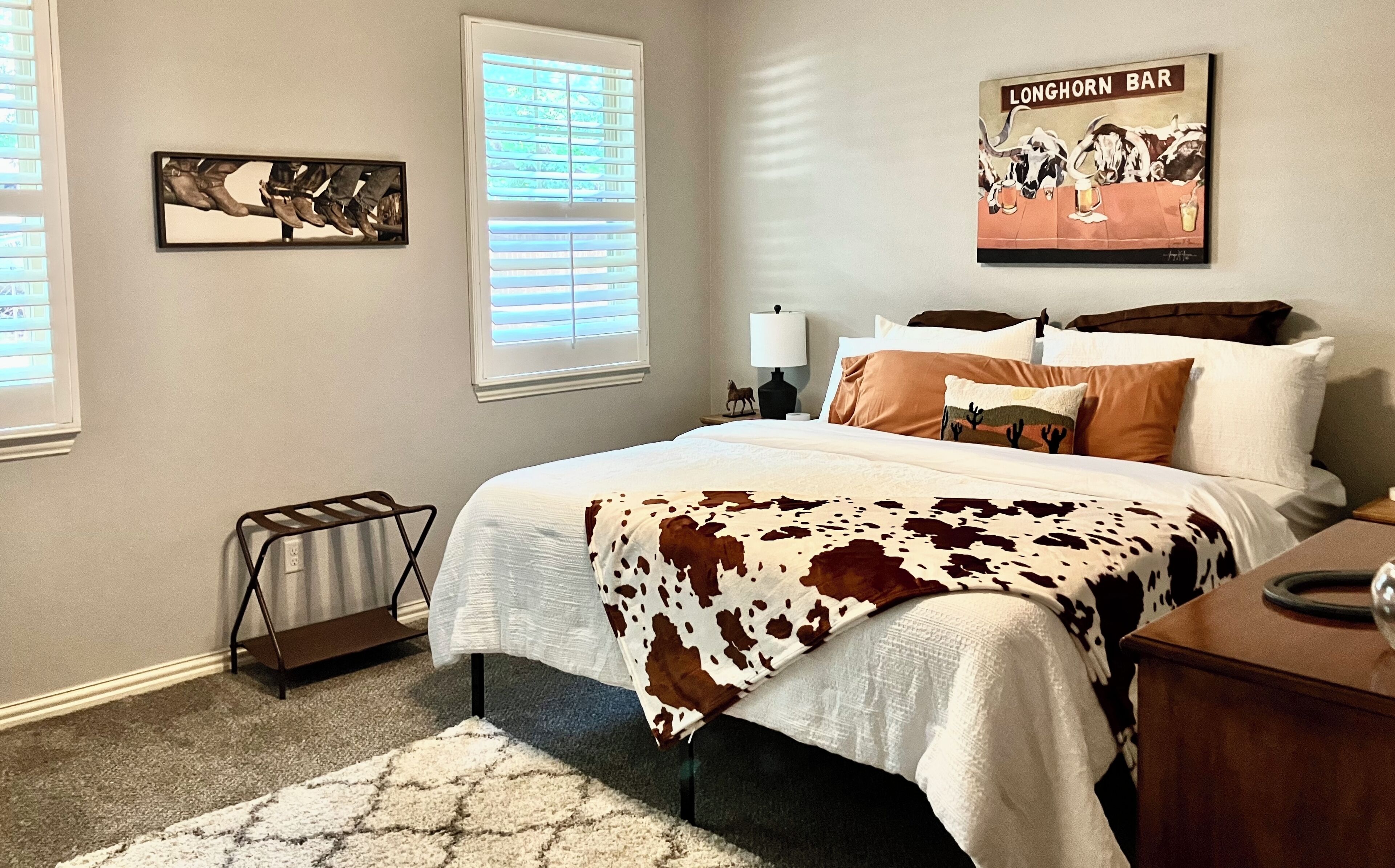 A bedroom with a cow print bedspread, a nightstand with a lamp, and a wall-mounted artwork depicting a bar scene.