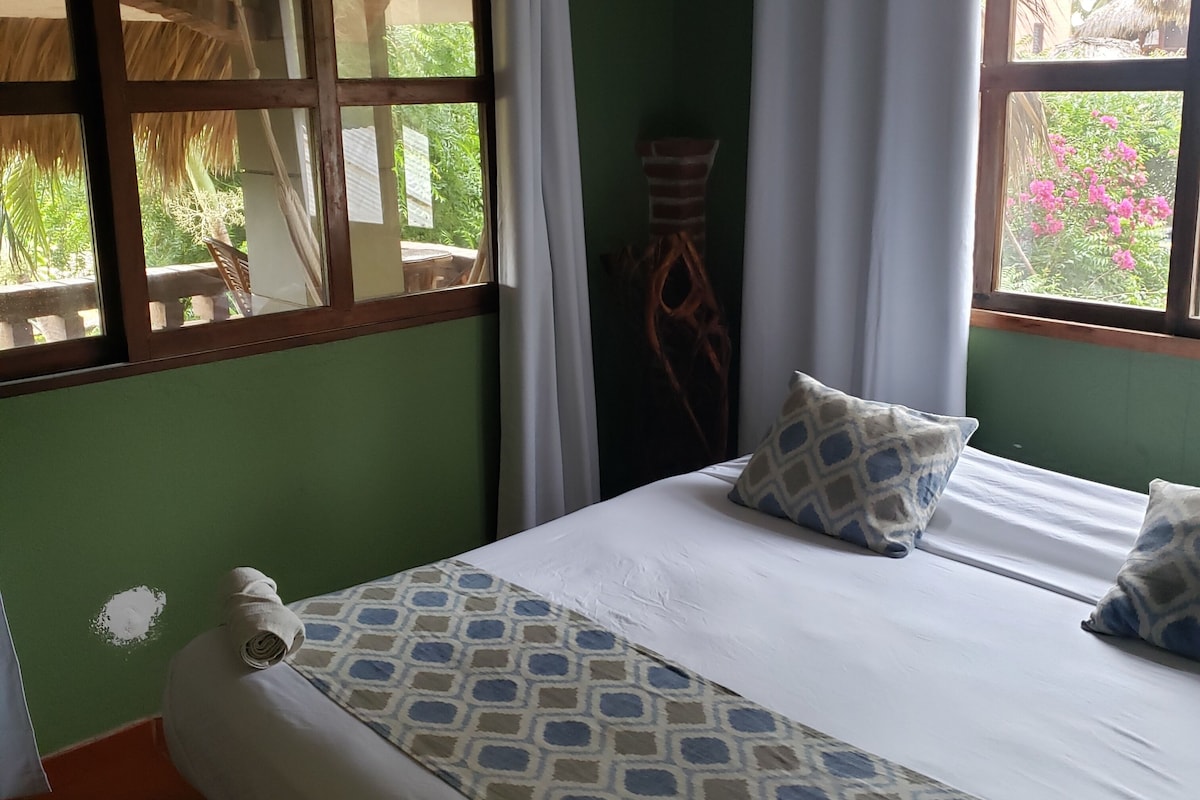 A room with a bed, two pillows, a window with curtains, and a view of greenery outside.
