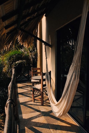 A hammock chair under a thatched roof, a wooden chair, and a glass door leading to a garden with palm trees. - Beachfrontguesthouse (Puerto Escondido)