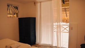 Une chambre à coucher avec un lit, une armoire de couleur sombre, une fenêtre donnant sur un bâtiment à toiture de chaume, et une porte menant à l’extérieur.