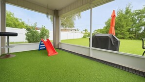 A covered patio area with artificial grass, a slide, and a table with an umbrella. - Located Midway Orlando & Daytona Beach, Pet Friendly, Fenced Yard, 2 Car Garage (Deltona)