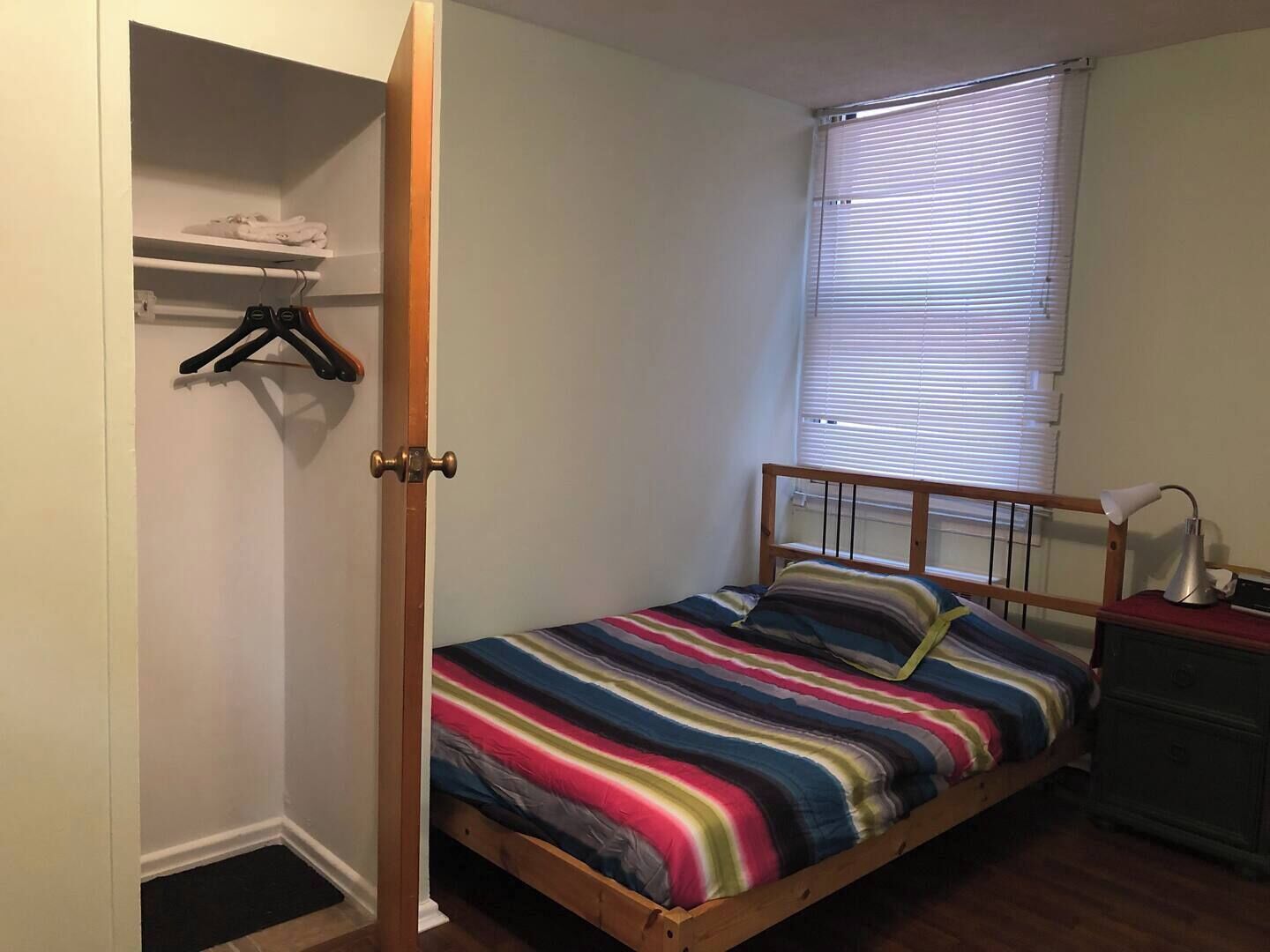 A single bed with a colorful striped bedspread, a wooden headboard, a bedside table with a lamp, and a window with blinds.