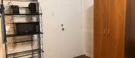 A room with a white door, a wooden wardrobe, and a metal shelf unit with kitchen appliances.