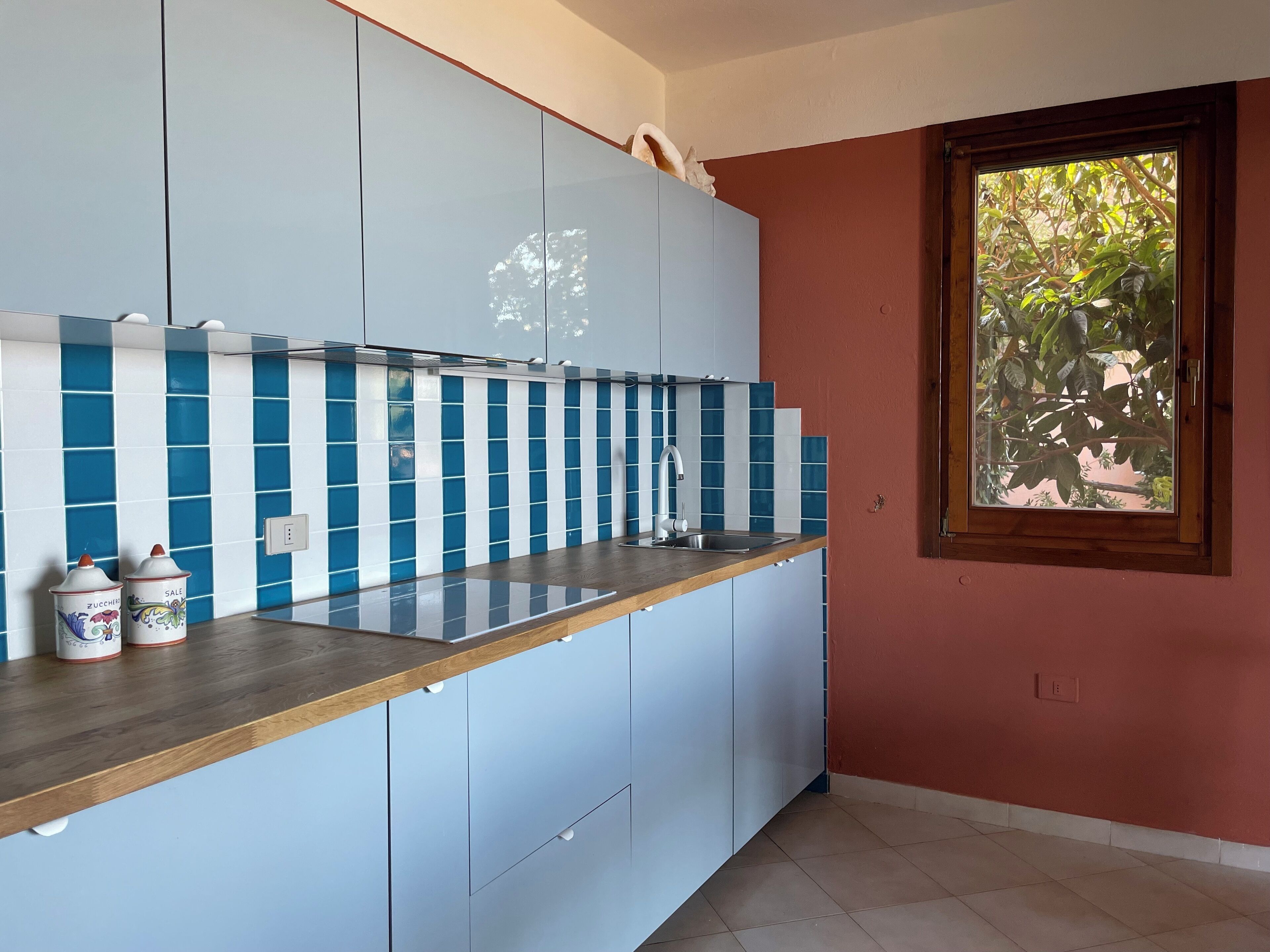 Une cuisine aux carreaux bleus et blancs, dotée d’un plan de travail en bois et d’une fenêtre donnant sur un paysage verdoyant.