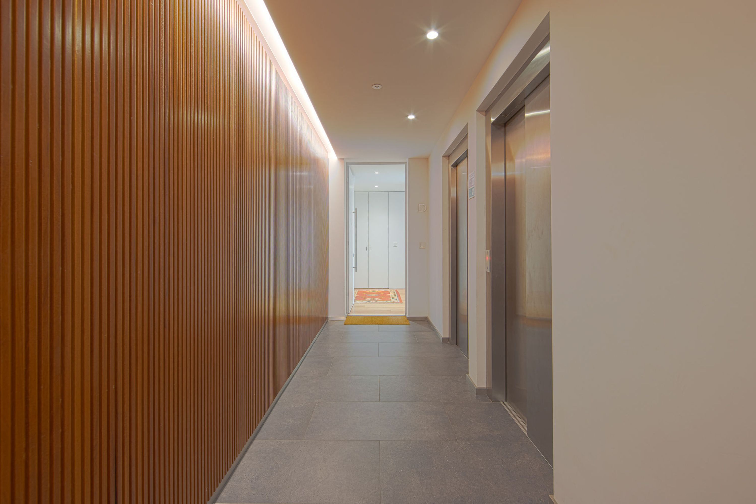 A modern hallway with wooden paneling on one side, a tiled floor, and an elevator on the right.