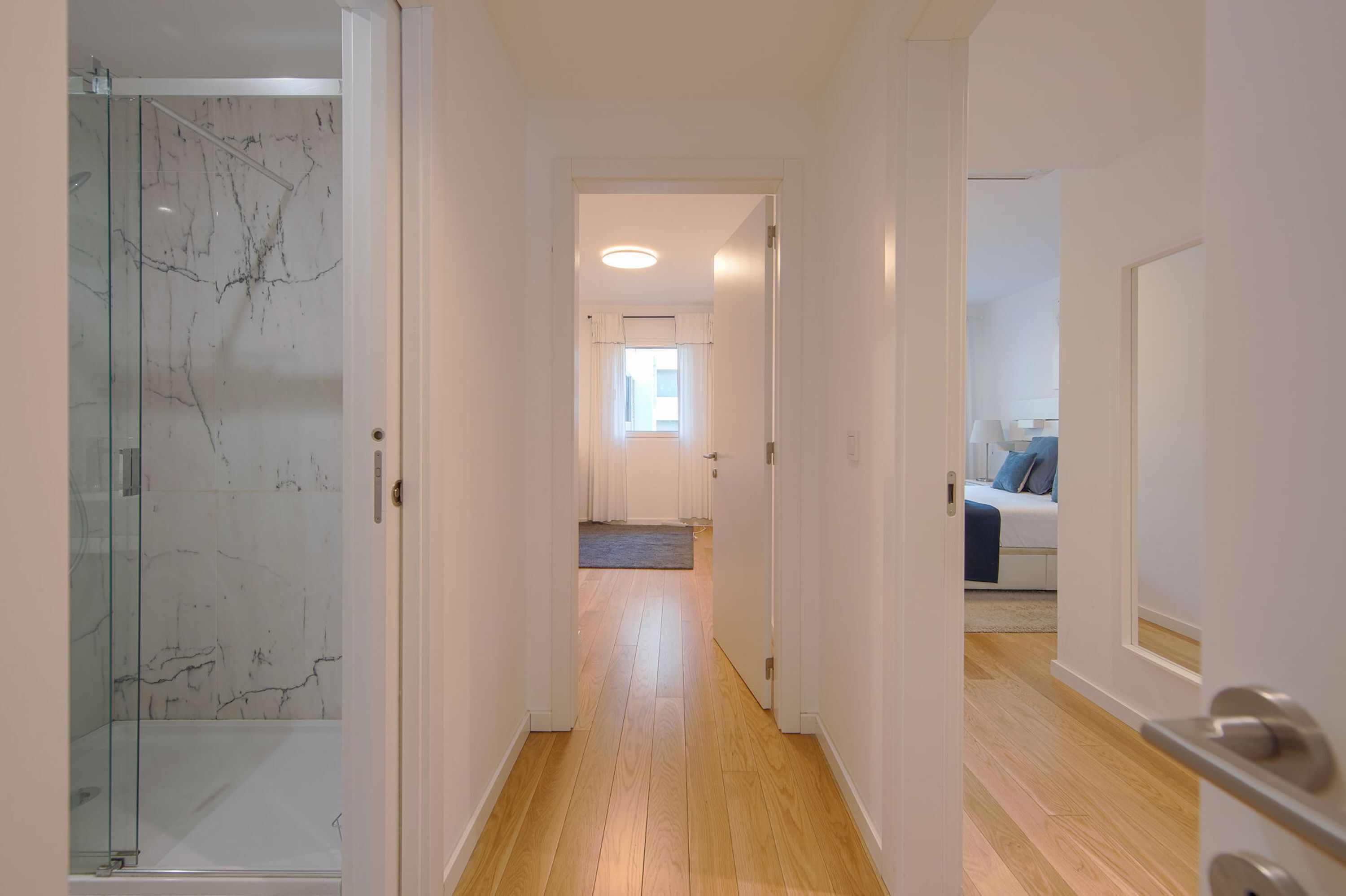 A hallway with a glass shower, wooden flooring, and a view into a bedroom with a bed and a window.
