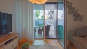 Ein modernes Wohnzimmer mit Flachbildfernseher, einer Couch, einem Esstisch mit Stühlen, einem großen Fenster mit Vorhängen und einem Balkon mit Blick auf Gebäude und Grünflächen.