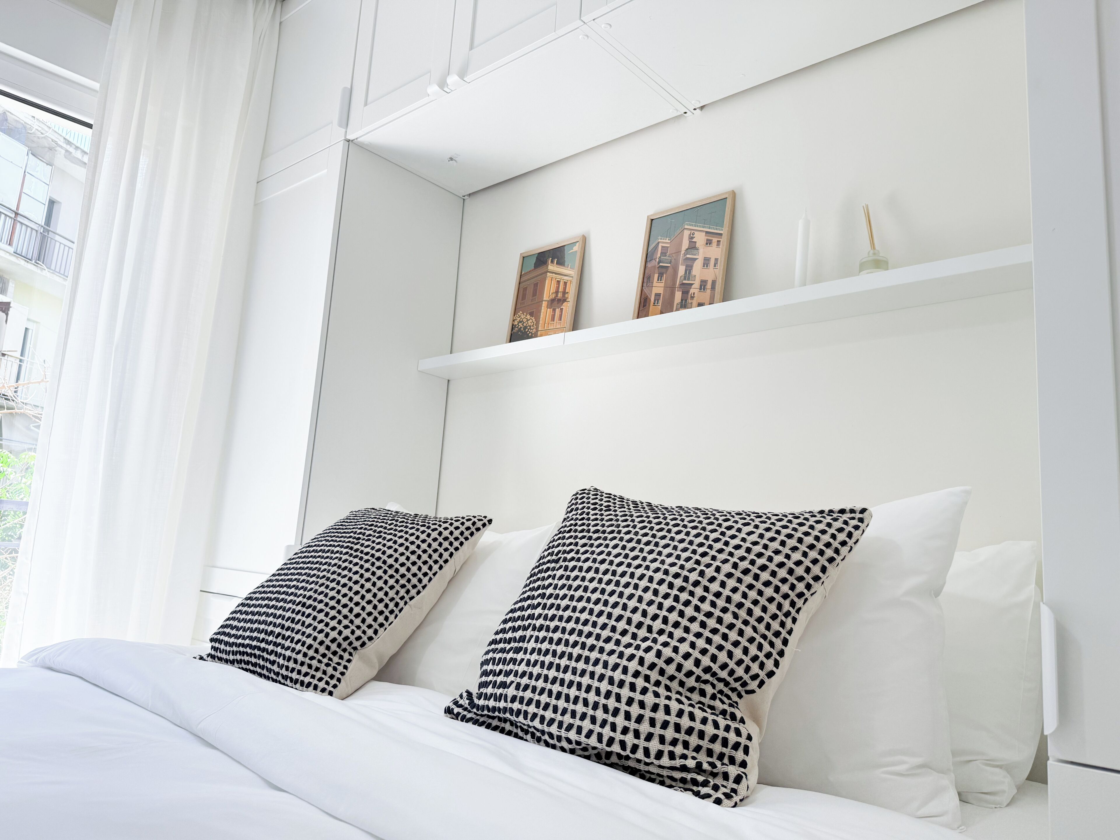 A hotel room with a bed, white bedding, two patterned pillows, a white nightstand, and a window with white curtains.