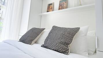 A hotel room with a bed, white bedding, two patterned pillows, a white nightstand, and a window with white curtains.