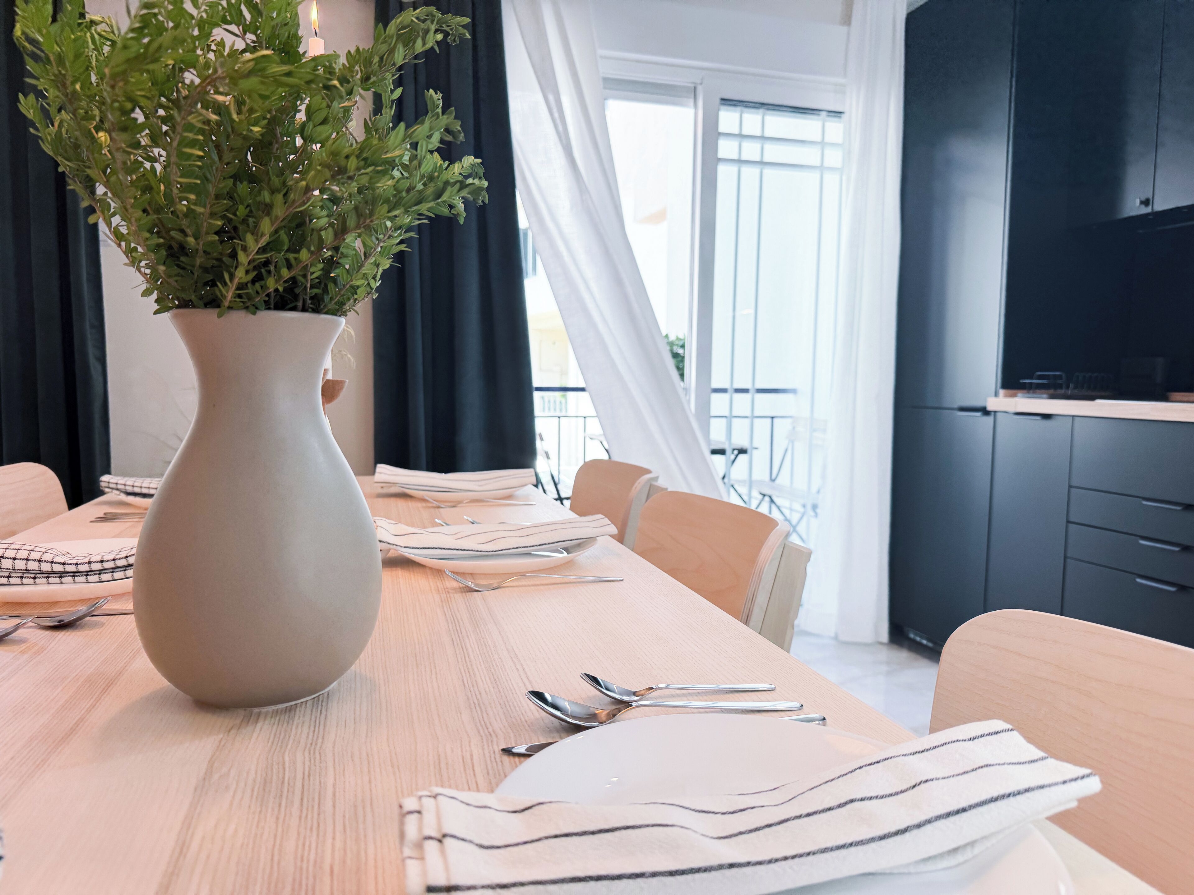 A dining table with a vase of green plants, neatly arranged cutlery, and striped napkins.
