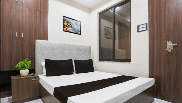 A bedroom with a bed, a nightstand, a potted plant, a window, and a painting on the wall. - Hotel S Grand  (Prayagraj)