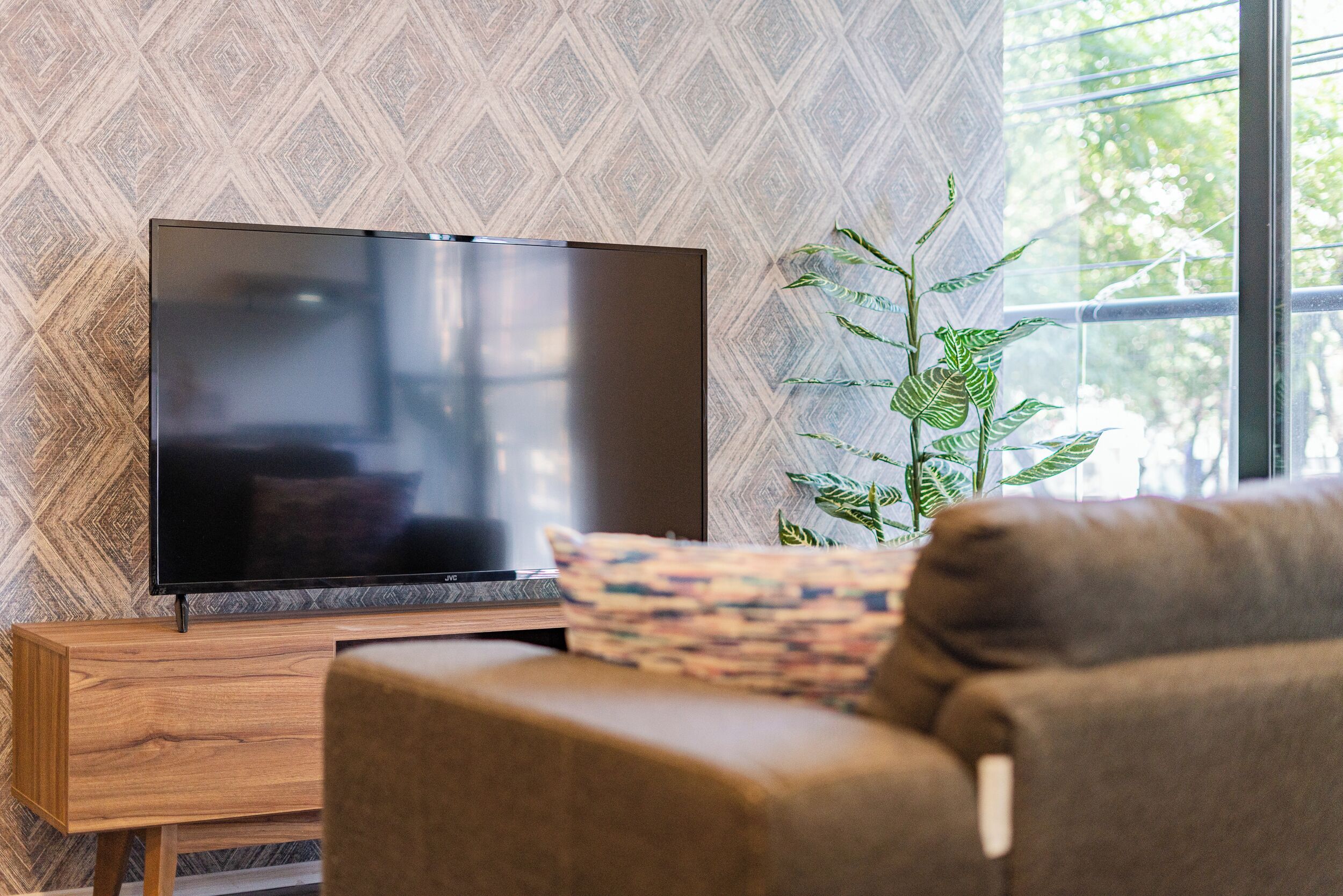 Ein modernes Wohnzimmer mit einem Flachbildfernseher auf einem Holzständer, einem braunen Sofa und einer Topfpflanze.