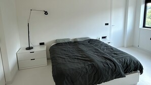 Une chambre à coucher au style minimaliste, avec un grand lit, une table de chevet, une lampe sur pied et une fenêtre.