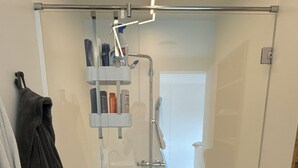 Une douche fermée par des parois en verre, dotée d’un pommeau de douche à main et d’un robinet mural. Des articles de toilette sont disposés sur une étagère au-dessus de la douche.