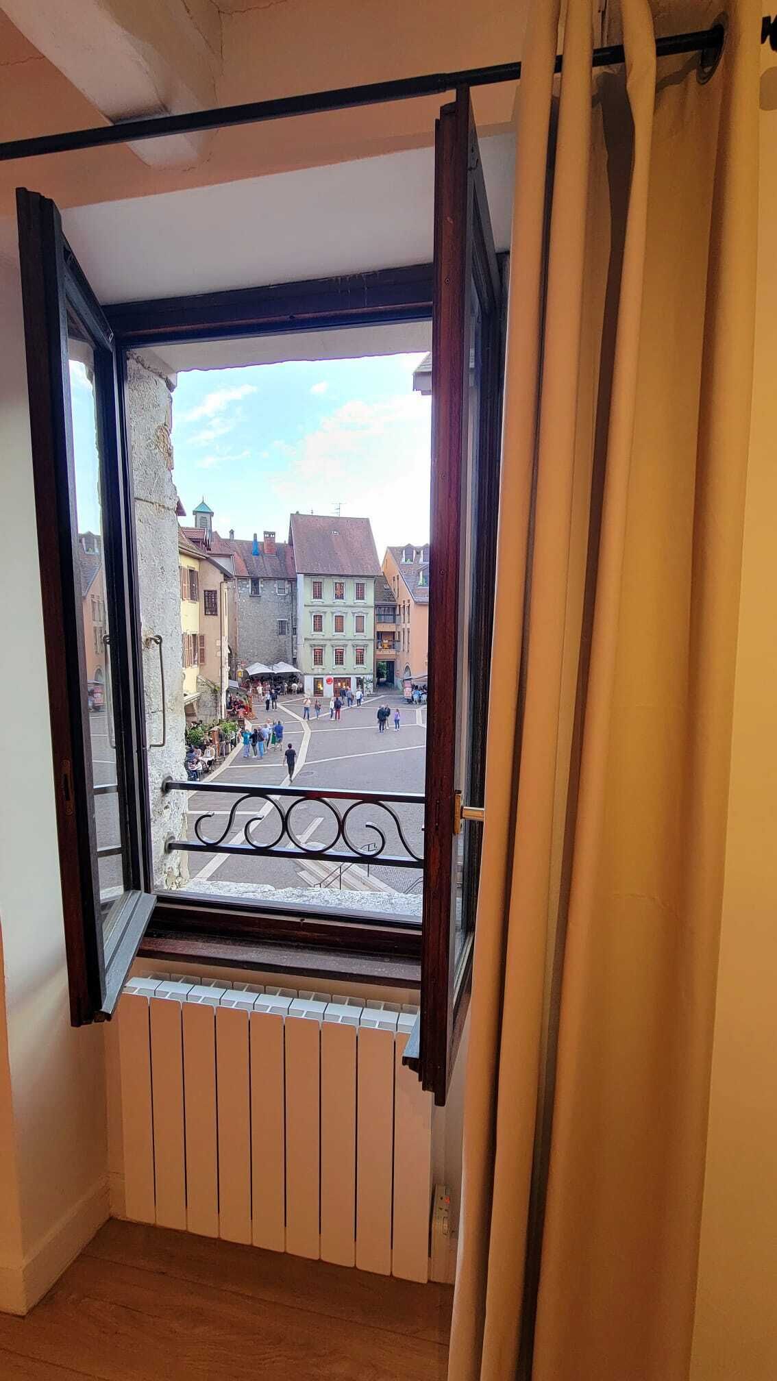 Et værelse med et vindue der kigger ud over en byplads, en radiator under vinduet, og et gardin halvt trukket fra.