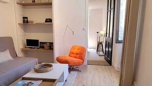 Ett modernt vardagsrum med en grå soffa, en orange fåtölj och ett soffbord. Det finns en bokhylla med diverse föremål och en lampa. Rummet har trägolv och ett stort fönster med gardiner.